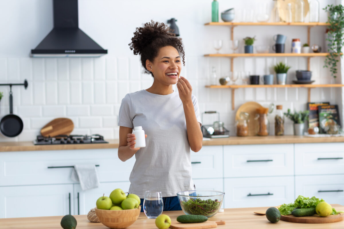 家庭のキッチンで料理台に立ち、サプリメントを飲む幸せなアフリカ系アメリカ人女性、遠くを見て親しげに微笑む、健康的なライフスタイルのコンセプト。