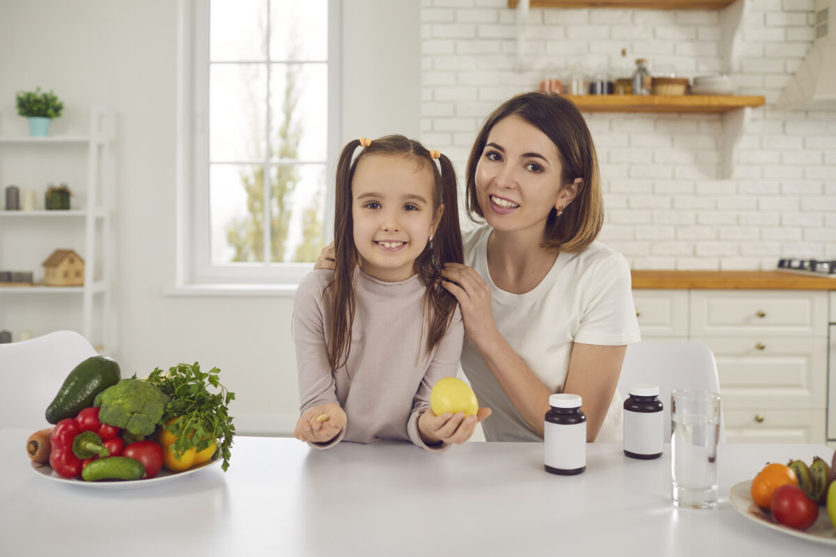 Porträtt av leende mamma och barn vid köksbordet med färsk frukt och grönsaker. Glad ung mamma och dotter äter hälsosam mat och tar C-vitamin och kosttillskott för att förebygga influensa och förkylning.